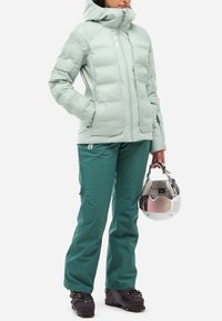 Helle mintgrüne Steppjacke mit Reißverschluss, Kapuze und strukturierten Einsätzen, kombiniert mit dunkelgrünen Skihosen und schwarzen Skistiefeln, während ein Helm gehalten wird.