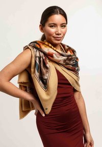 Wrap in beige fabric with fringe, layered over a reddish dress. The scarf features colorful abstract patterns in earthy tones.