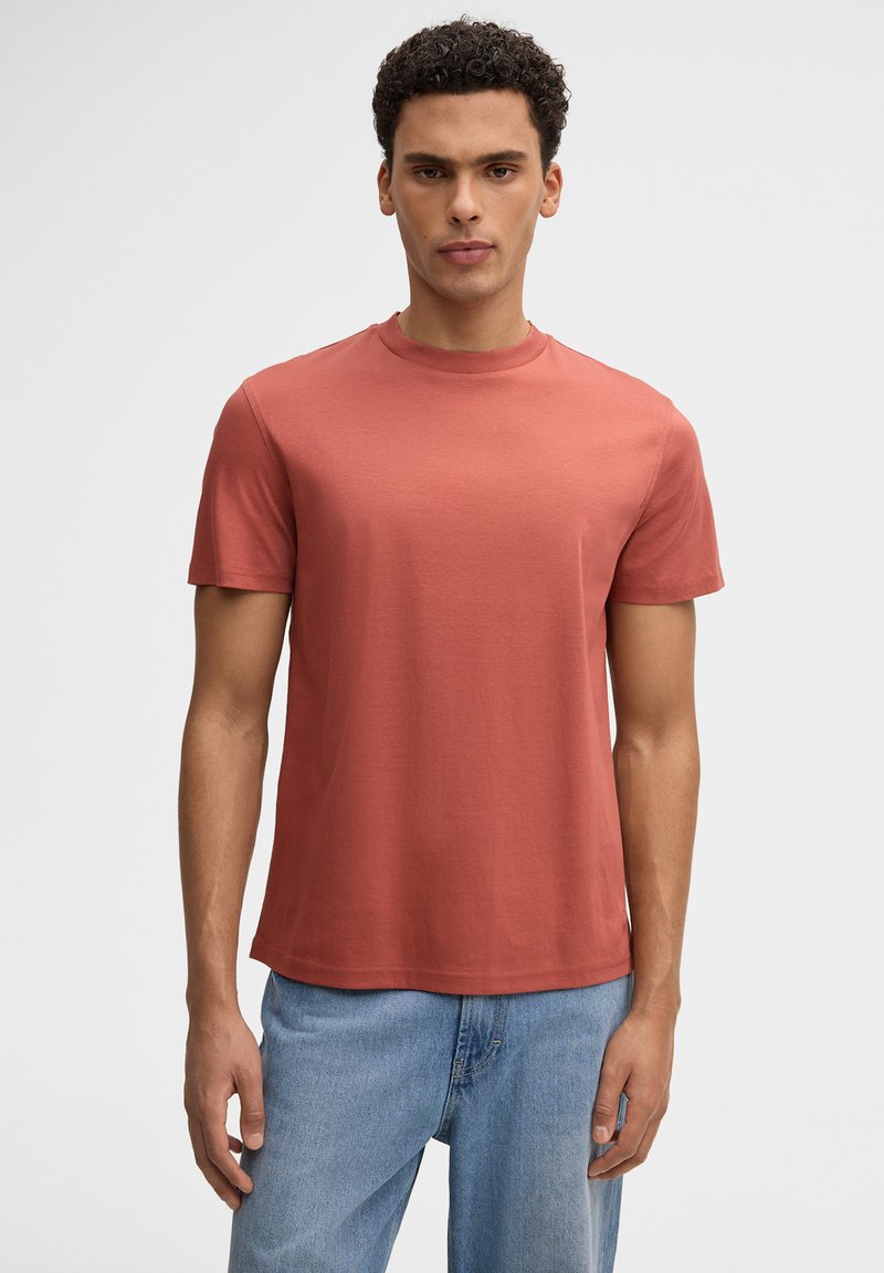 Junger Mann mit kurzen lockigen Haaren, der ein einfarbiges, rostfarbenes T-Shirt mit kurzen Ärmeln und hellblaue Jeans trägt und vor einem einfarbigen Hintergrund steht.