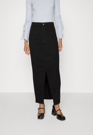 Frau trägt ein hellblaues Langarmoberteil und einen hoch taillierten schwarzen Midirock mit Schlitz vorne, kombiniert mit schwarzen Mary Jane Pumps.