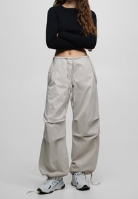 Person i beiga vida byxor med dragsko, vita sneakers och en svart långärmad cropped topp, står med armarna i kors mot en grå bakgrund.