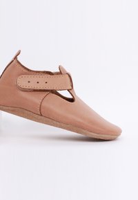 Miękkie, beżowe skórzane buciki dla niemowląt z paskiem T i zapięciem na rzep, przedstawione w profilu bocznym na białym tle.