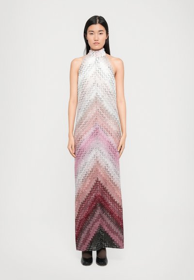 Kobieta stoi przodem, ubrana w bezrękawnikową sukienkę w gradient w stylu halter z wzorem chevron w kolorach białym, różowym, czerwonym i czarnym, w połączeniu z czarnymi butami.