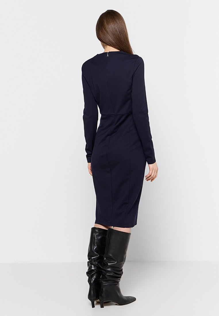 Donna con capelli castani lisci, che indossa un vestito blu scuro a maniche lunghe aderente e stivali neri con tacco al ginocchio, di spalle contro uno sfondo bianco.