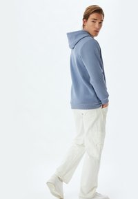 Sudadera azul con cordón y bolsillo tipo canguro, combinada con pantalones cargo blancos. El modelo está de lado, con las manos en los bolsillos, vistiendo zapatos blancos.