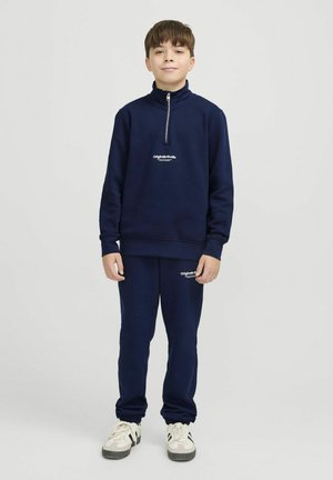 Sweatshirt zippé bleu marine avec col haut et logo sur la poitrine, associé à un pantalon de jogging fuselé assorti et des baskets de couleur claire.