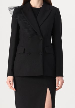 Femme portant un blazer noir croisé avec un ornement en tulle sur une épaule et une jupe noire fendue jusqu'en haut de la cuisse.