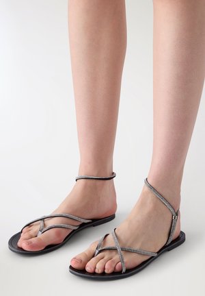 Piedi nudi che indossano sandali neri piatti con sottili cinturini argento scintillanti che attraversano le dita e circondano le caviglie su uno sfondo chiaro.