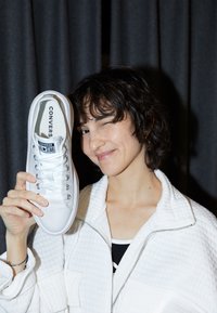 White canvas Converse All Star sneaker with lace-up front and metal eyelets, held by a person in a quilted white jacket against a dark backdrop.