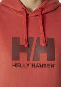 Červená mikina s kapucí z měkké tkaniny, která má velké hnědé logo "HH" a pod ním text "HELLY HANSEN" ve shodné hnědé barvě. Šňůrky na kapuci.