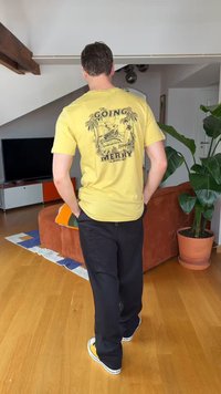 Homme portant un t-shirt graphique jaune « The Going Merry », un pantalon noir et des baskets jaunes, marchant dans un salon moderne avec un sol en bois et des plantes.