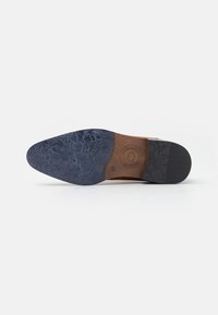 Semelle de chaussure avec une tige en cuir marron et une section en caoutchouc noir texturé, présentant un motif unique et un détail délicat de logo embossé.