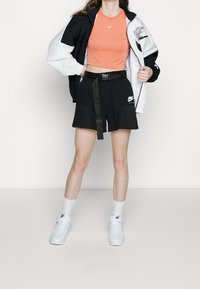 Žena v černo-bílé bundě Nike, oranžovém crop topu, černých šortkách Nike s páskem, bílých ponožkách a bílých teniskách Nike.