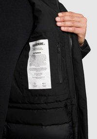 Black insulated jacket featuring a soft texture, zippered pocket, and a label on the inner lining with care instructions and branding.