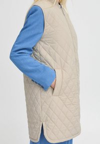 Beige gesteppte Weste mit hohem Kragen und Fronttaschen, kombiniert mit einem blauen Langarmhemd mit knöpfbaren Manschetten.