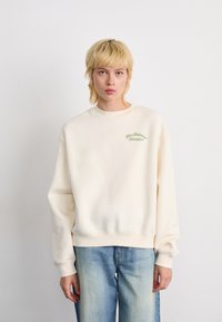 Persoon met kort blond haar, gekleed in een oversized crèmekleurig sweatshirt met groene tekst "New Balance Athletics" en lichtblauwe vervaagde spijkerbroek.