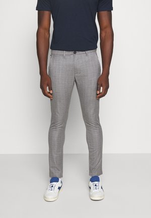 Mężczyzna ubrany w szare spodnie slim fit, białe sneakersy z niebieskimi akcentami oraz ciemnogranatową koszulę z krótkim rękawem, stojący na jednolitym tle.