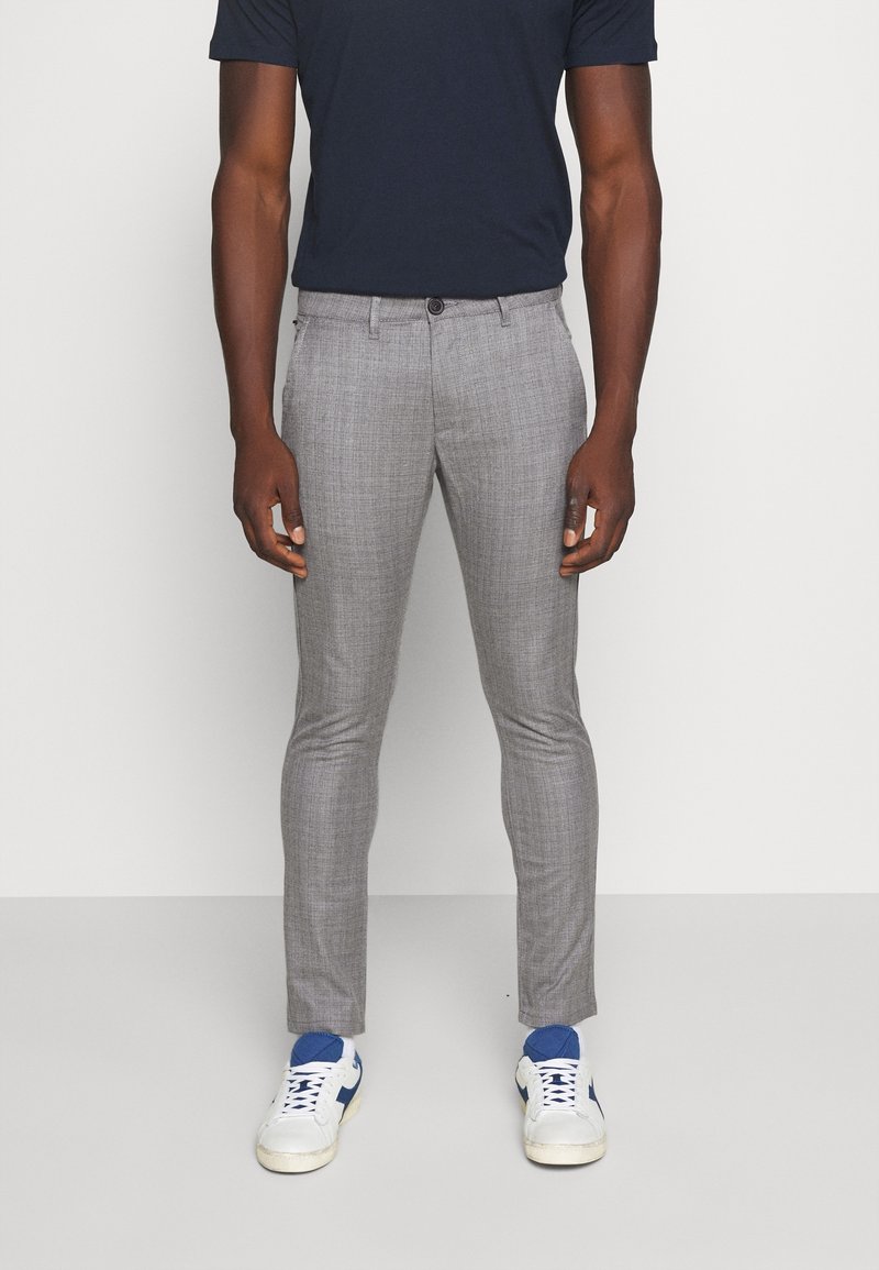 Homme portant un pantalon gris slim, des baskets blanches avec des accents bleus et une chemise à manches courtes bleu marine foncé, debout devant un fond uni.