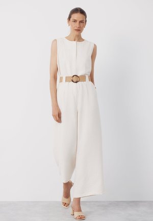 Frau trägt weißen ärmellosen Jumpsuit mit weitem Bein, beigen Gürtel mit runder Schnalle und beige Sandaletten mit Absatz, steht vor weißem Hintergrund.