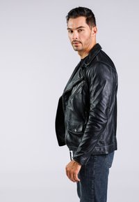 Giacca da motociclista in pelle nera con cerniera asimmetrica, tasche laterali e finitura texturizzata, indossata sopra una maglia nera, mostrata su uno sfondo neutro.