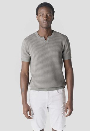 Hombre con camisa de punto de manga corta de color gris claro y pantalones cortos blancos rasgados, de pie contra un fondo liso de color gris claro.