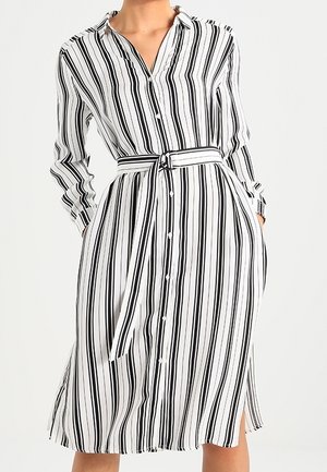 Vrouw die een witte midi-jurk draagt met verticale zwarte strepen, lange mouwen, knopen aan de voorkant, zijschotten en een bijpassende ceintuur in de taille.