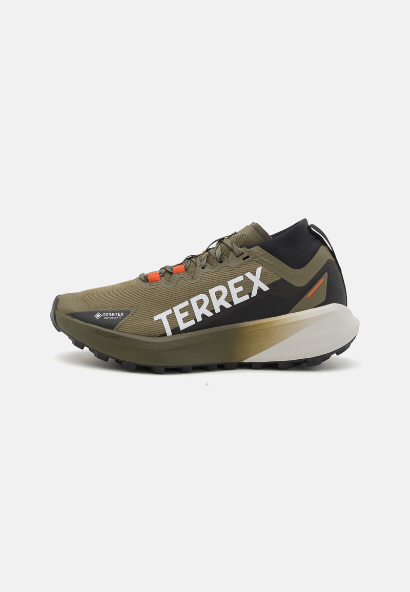 Zaļas un melnas taku skriešanas kurpes ar strukturētu augšdaļu, lielu baltu 'TERREX' logo, oranžiem akcentiem un biezu, izturīgu zolu.