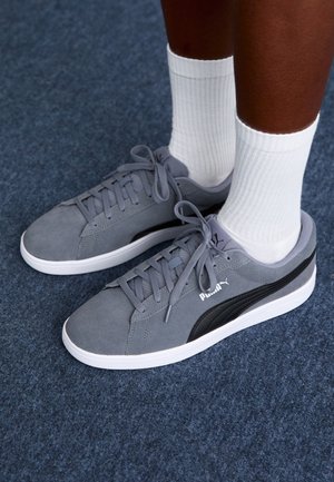 Gray suede sneakers with black accents, white rubber sole, and round laces. Paired with ribbed white socks. Background features navy carpet.