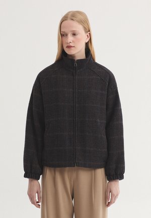 Young woman with straight blond hair wearing a dark checkered zip-up jacket and beige pants, standing against a plain light background.