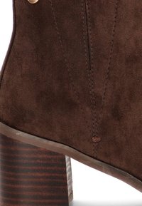 Botín de ante marrón con una superficie texturizada, detalles de costura vertical y un tacón de madera grueso y a rayas. Acentuado con herrajes dorados.
