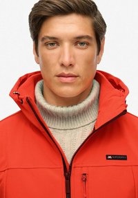 Veste à capuche rouge avec fermeture éclair, en tissu texturé, et logo sur la poitrine, superposée sur un pull à col roulé en tricot gris clair.