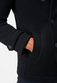 Schwarze Jacke aus strukturiertem, gestricktem Stoff. Verfügt über knöpfbare Manschetten und eine Seitentasche. Das Design ist strukturiert mit einer klaren Silhouette.