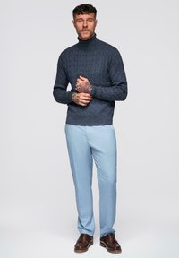 Pull à col roulé en tricot torsadé bleu marine associé à un pantalon bleu clair et des chaussures marron. Le modèle se tient les mains jointes devant.