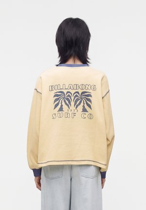 Persona con capelli scuri fino alle spalle indossa una felpa beige Billabong Surf Co con motivo di palme, abbinata a jeans azzurri chiari, rivolta di spalle.