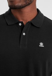 Black polo shirt featuring a textured fabric, two white buttons, and a small embroidered logo on the left chest.