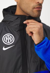 Sort jakke med høj krave, lynlås-lukning, fleece-blå ærmer, Nike og logo patches på brystet. Glat, vandafvisende materiale.