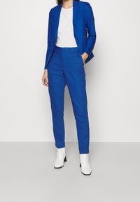 Kvinna klädd i en ljusblå skräddarsydd kostym med smala byxor och en kavaj, matchat med vita ankelskängor och en vit topp.