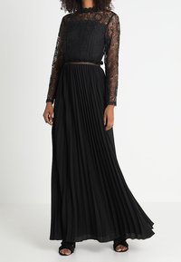 Donna che indossa un lungo abito nero con un top in pizzo e una gonna plissettata, abbinato a sandali neri con punta aperta.