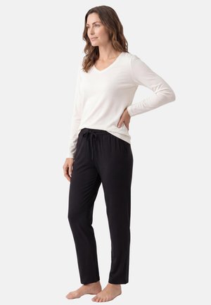 Donna in piedi scalza, indossa una maglia bianca a maniche lunghe e pantaloni neri con coulisse su uno sfondo chiaro e uniforme.