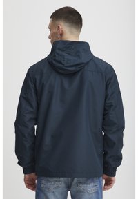 Marineblå vindjakke med hette, med elastiske mansjetter og strukturert stoff. Bakdelen har et enkelt design uten logoer.