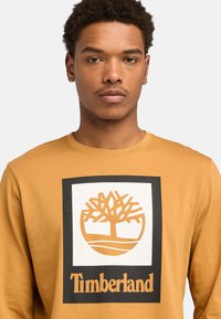 Long-sleeve, mustard-colored T-shirt featuring a black-framed white logo with a tree and the word "Timberland" in bold lettering.