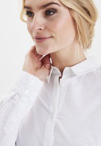 Camicia bianca button-up con maniche lunghe e colletto a punta. Il tessuto appare liscio con una sottile trama e dettagli di bottoni visibili sui polsini.