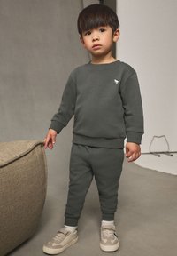 Dunkelgrüner Pullover und Jogginghose aus weichem Stoff, mit einem kleinen weißen Logo auf der Brust, kombiniert mit beigen Sneakern und weißen Socken.