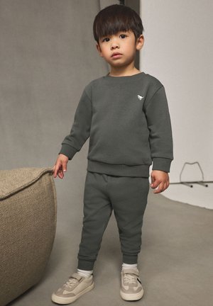 Sweat-shirt vert foncé et pantalon de jogging en tissu doux, avec un petit logo blanc sur la poitrine, accompagné de baskets beiges et de chaussettes blanches.