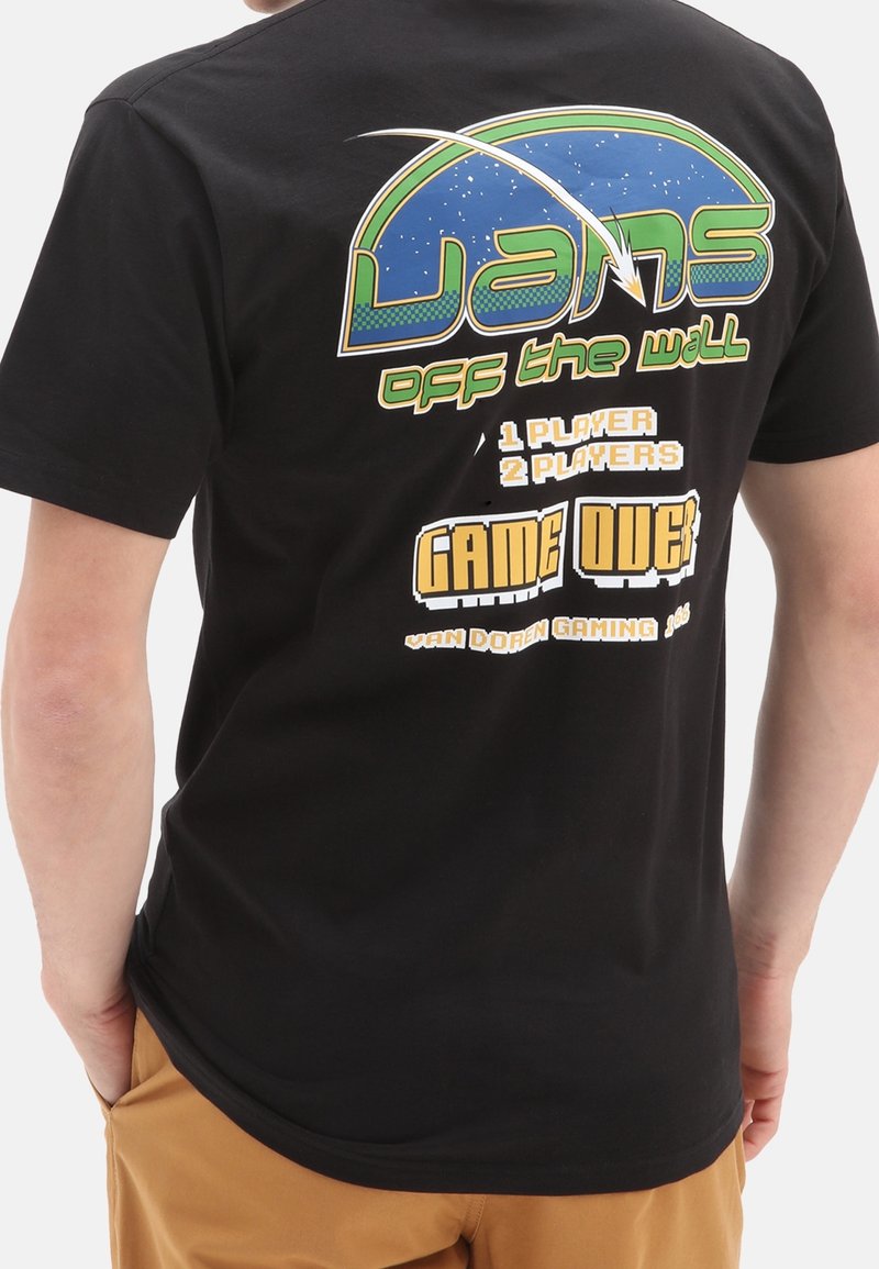 Personne portant un T-shirt noir Vans avec un design rétro gaming et le texte "1 player 2 players Game Over" dans le dos.