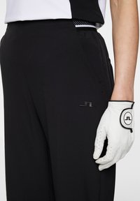 Svarta golfbyxor med en slät tygkvalitet, som har sidfickor och en diskret logotyp på vänster lår. En vit golfhandske kompletterar outfiten.