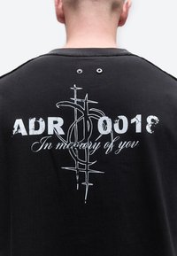 Persoon draagt een zwart shirt met witte tekst "ADR 0018 Ter nagedachtenis aan jou" en een gestileerd kruisontwerp op de achterkant.