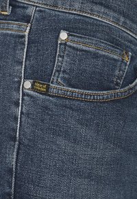 Mörkblå denimjeans med en klassisk femfickdesign, gula sömmar och en "Tiger of Sweden"-etikett på bakfickan.