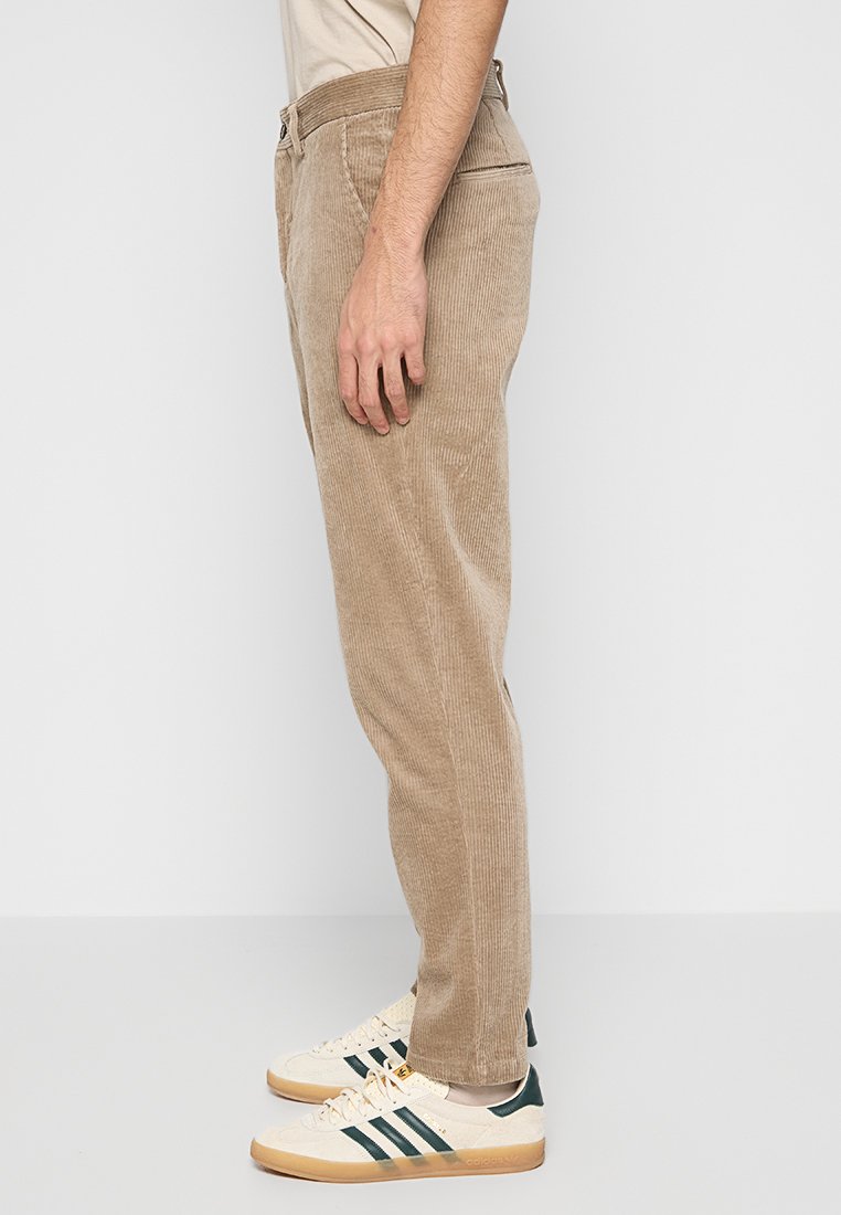 Beige Corduroyhosen mit geradem Bein, die eine strukturierte Oberfläche und Seitentaschen aufweisen. Kombiniert mit cremefarbenen Turnschuhen und grünen Akzenten.