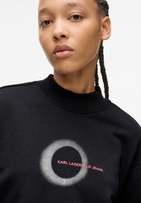 Fekete pulóver magas gallérral, kerek fehér grafikával és rózsaszín "KARL LAGERFELD JEANS" felirattal. Puha tapintású.